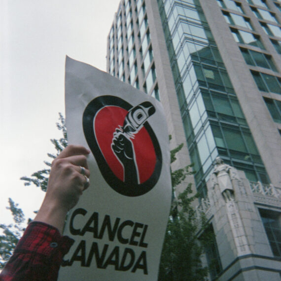 Photo of a person’s arm holding a sign in the air that says “Cancel Canada”. The sign features an illustration of a hand holding a feather with a formline design within it. The hand is framed by a solid red circle outlined by a thick black circle. The person is wearing a red and black plaid shirt. There is a tall building and traffic lights behind the sign.