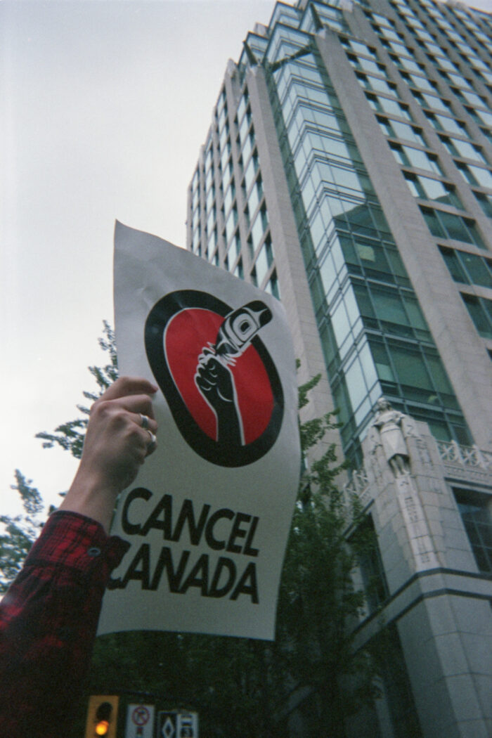 Photo of a person’s arm holding a sign in the air that says “Cancel Canada”. The sign features an illustration of a hand holding a feather with a formline design within it. The hand is framed by a solid red circle outlined by a thick black circle. The person is wearing a red and black plaid shirt. There is a tall building and traffic lights behind the sign.