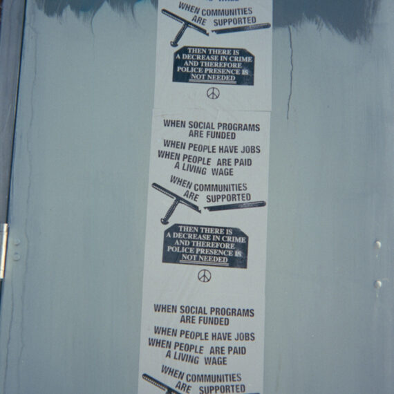 Three of the same poster wheat pasted on a grey-blue door. The poster reads “When social programs are funded, when people have jobs, when people are paid a living wage, when communities are supported, then there is a decrease in crime and therefore police presence is not needed.” The poster has an illustration of a broken police club above a black prism. Underneath the prism is a peace symbol..