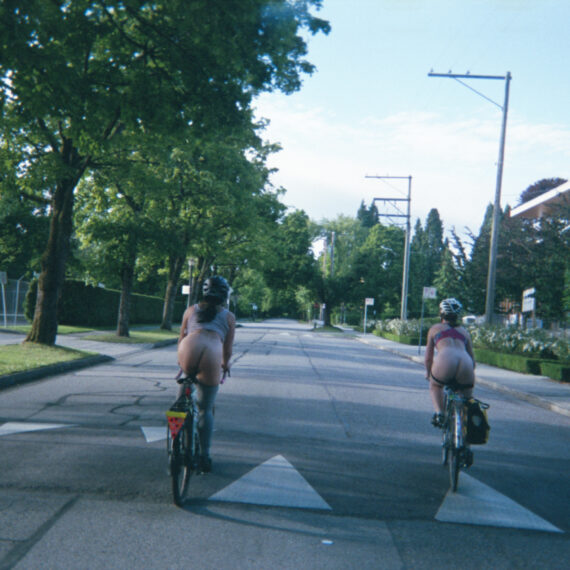 Two pantless people wearing sports bras and helmets are riding bikes along a residential street. They are riding over a speed bump. On either side of the street are shrubs and trees. The sky in the distance is pale blue with thin clouds scattered across it.