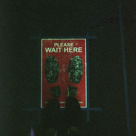 Photo of a vinyl sign on the floor that reads “Please wait here.” The sign is red with two black foot prints. The photographer is looking down at their shoes which are stepping partially on the sign.