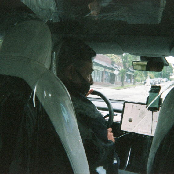 Photo taken from the backseat of a car. The driver is wearing their face mask below their nose while looking to the right. In front of them is a phone attached to a stand and the navigation system screen of the car. The interior of the car is wrapped in plastic.