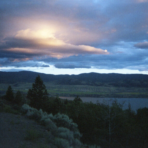 Photo of a landscape with the sun shining through the clouds. The clouds surrounding the sun are glowing while the other clouds are a light blue-grey. Beneath the sky are rolling mountains, grassy fields, evergreen trees, and a body of water.
