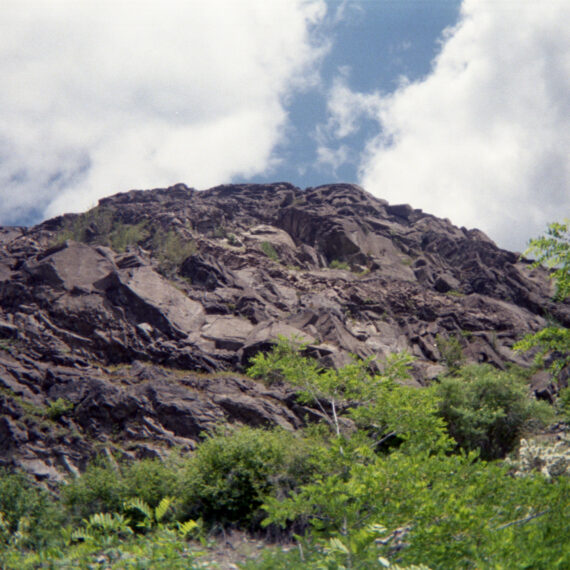 Photo of a rocky hill in front of a blue sky filled with large clouds on either side. In front of the rocky hill are small trees and shrubs.