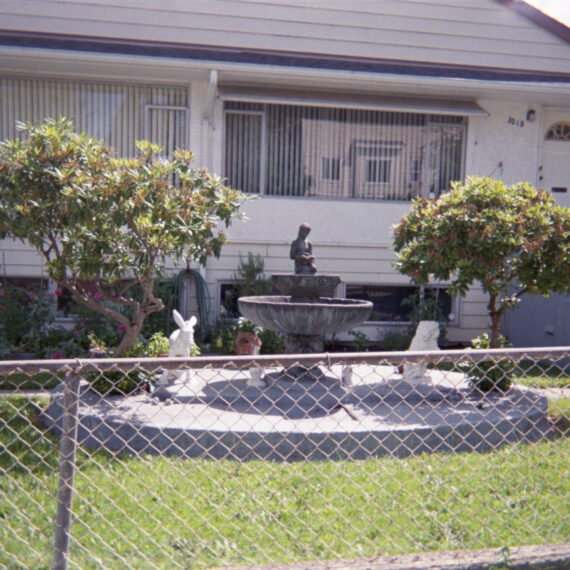 Photo of a front yard of a house behind a wired fence. There is a stone fountain with sculptures and shrubs surrounding it. The house is white with two large windows facing the street.