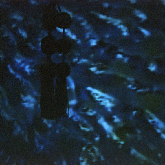 Photo of a projection of water against a wall. There is a string of three knots hanging on the wall of the projection. The photo is dark and underexposed.