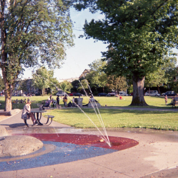 Photo of a water park feature at a neighbourhood park. There is a person sitting at black bench and table behind the water park. In the distance are more people gathering on the grass in between two large trees. The sky is bright and blue.