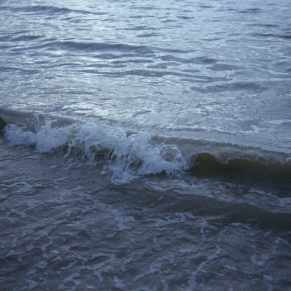 A small wave crashing against a larger body of water. There is water being illuminated by light in the distance.