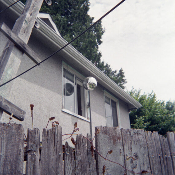 A facade of a house surrounded by a jagged wooden fence. There is a dried vine growing between the gaps of the fence. A side window of the house is open with a disco ball hanging outside of it. Behind the house are tall evergreen trees and a cloudy blue-grey sky.