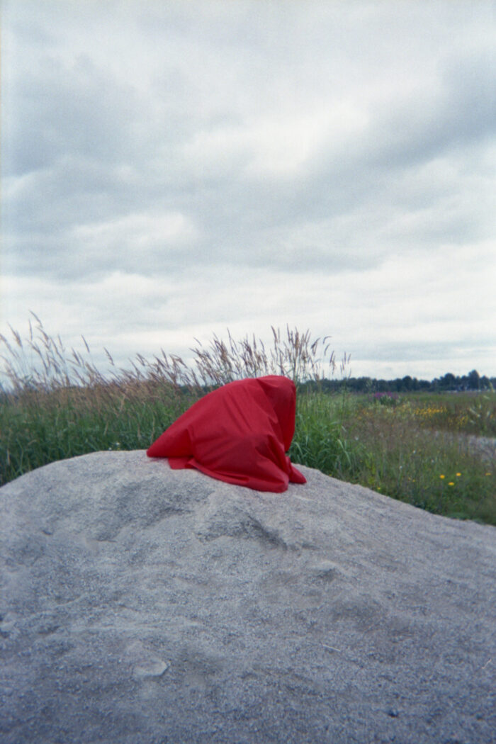 A person completely covered in red cloth is kneeling on a pile of gravel in front of a field of long grasses. Sky is cloudy in the background.