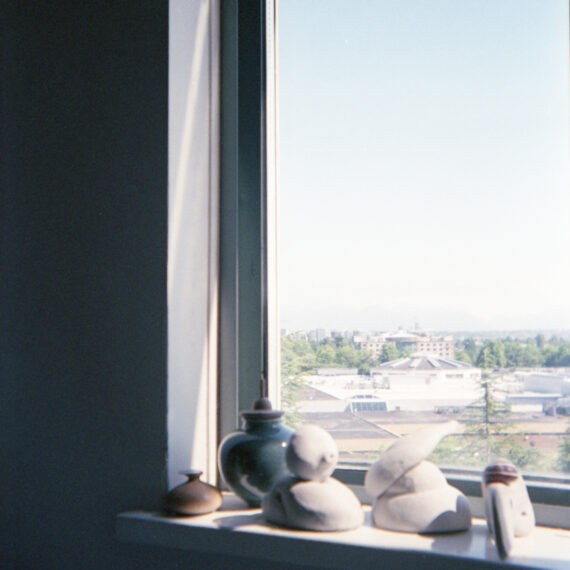 Photo of a window sill lined with ceramic pots of various sizes. The window is looking out to the rooftops of various buildings. The faint silhouettes of mountains can be seen along the horizon of a pale blue sky.