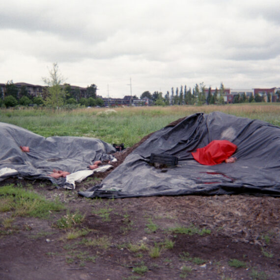 A red cloth covering something is resting on a black tarp covering a pile of dirt. There is a smaller pile of dirt covered in black tarp adjacent to the first piel. Behind the piles of dirt is a grassy area and residential buildings in the distance. The sky is cloudy.