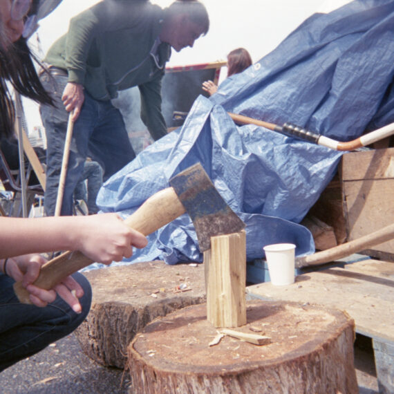 An Indonesian person with long black hair wearing glasses is chopping firewood on a stump using an ax. Behind them is a white person with short hair wearing a green sweater and jeans holding a stick and leaning towards a pile of wood covered by a blue tarp. Smoke is rising in the background.