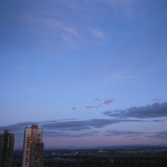 View of a skyline with high rise buildings scattered across the horizon. The sun is setting and the sky is a blue-purple colour with a wash of pink at the lowest point of the horizon. There are elongated clouds spread out across the sky.
