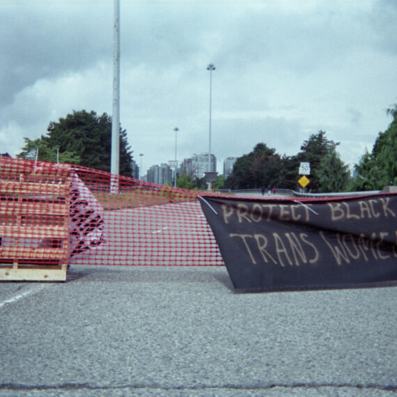 A viaduct in Vancouver’s downtown is blocked off with red plastic netting attached to a wooden pallet. There is a black cloth zip-tied to the red netting spray painted in gold with the text “Protect Black trans women.” In the distance are high rise buildings, street lamps, and evergreen trees.