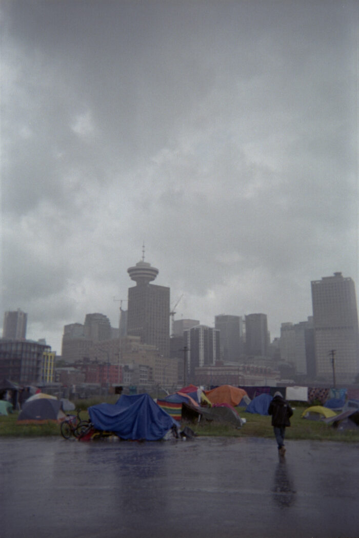 A person wearing a black jacket is walking towards a cluster of tents set up along a fence and on the grass. It is a tent city. In the distant background are high rise buildings with the iconic Harbour Centre visible along Vancouver’s city skyline. The sky is grey with overcast and completely covered with rain clouds.