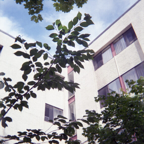Photo of an apartment complex with a white stucco facade surrounded by trees. There are open and closed windows with curtains drawn facing one another. In the background is a pale blue sky with faint fluffy clouds.