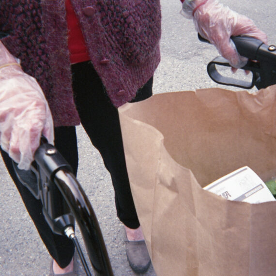 A senior wearing a purple button-up sweater, red shirt, black pants, grey shoes, and plastic gloves is using a black rollator. Only the bottom half of their torso can be seen. On the walker is a paper bag filled with groceries and a piece of paper.
