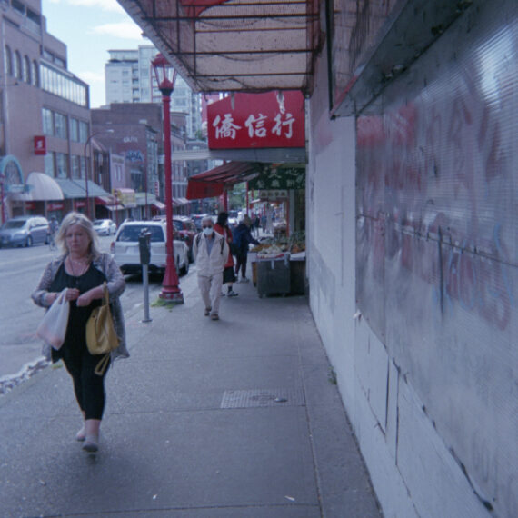 Photo of a pedestrian with long silver hair wearing a black shirt and pants carrying two bags walking towards the camera. Behind them is another person wearing a face mask, sunglasses, and a cargo vest walking towards the camera. The awnings of shops with Chinese text on them along the block can be seen in the distance on the right. Red street lamps line the streets.