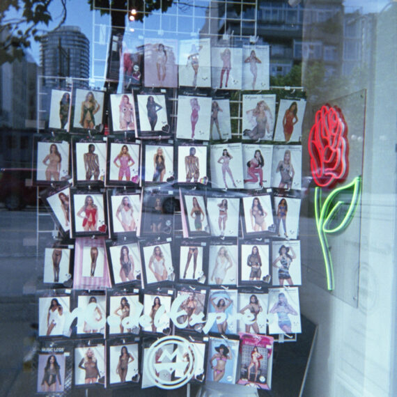 A window display in a retail store. There is a white steel rack holding packages of clothing with photos of women on the front. There is a neon light installation shaped like a red rose next to the rack of products. There is white vinyl lettering on the window that reads “Model Express” with the store logo underneath.