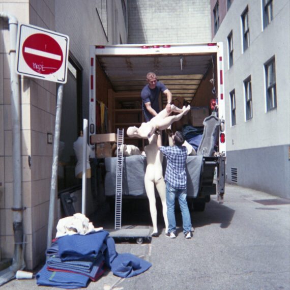 Two people unloading a truck full of mannequins in an alleyway. There is one person with short hair wearing a navy blue shirt passing a mannequin without arms to a person with short hair wearing a plaid shirt and jeans.