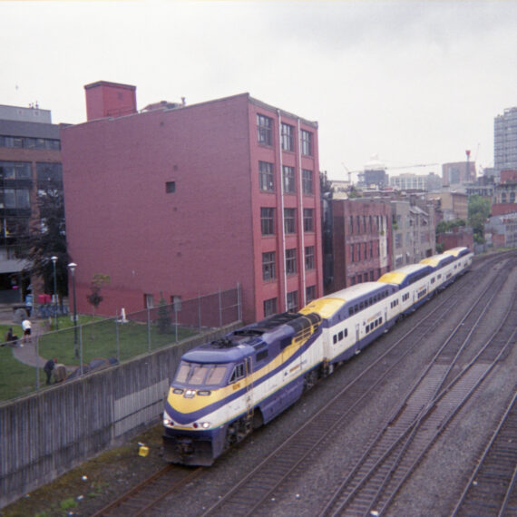 A blue, yellow, and white train on the train tracks is stopped next to a red building. Next to the front of the train is a concrete wall and metal fence. Behind the fence is a grass area where people are sitting and resting.