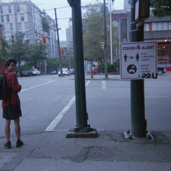 A cross walk with a person wearing a red shirt and black shorts waiting to cross the street. Next to him is a sign attached to a green pole that reads “COVID-19 Alert. Stay 2 metres apart. Wash your hands”. There is some graffitied text on the sign in black that reads “Do U”. The person standing at the crosswalk is approximately 1 metre away from the sign and they are staring back at the camera.