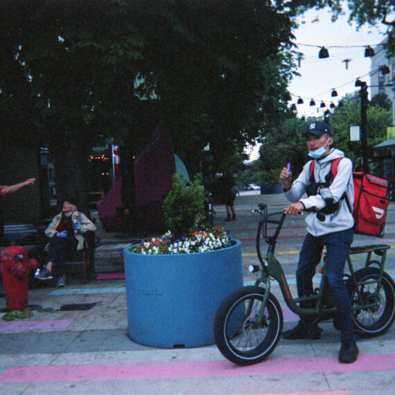 A person riding a small green bike is wearing a white hoodie, blue jeans, black sneakers, a navy blue baseball cap, and a bright red DoorDash food delivery backpack. Their blue surgical face mask is pulled down to their chin and they’re holding out a thumbs up. In the background are two people conversing by a bench, trees, a fire hydrant and crossing pedestrians.