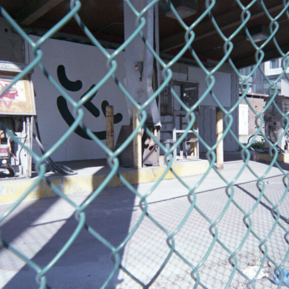 A graffitied smiley face in black paint on a white wall. The photo shows the wall through a green wired fence. Next to the wall are abandoned gas pumps on a concrete block.