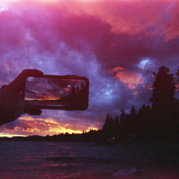 A sunset sky at a beach shore. Black silhouettes of evergreen trees can be seen across the horizon. The sky is a brilliant pink orange with dark blue clouds sweeping across the sky. The water reflects the colour of the. A hand is holding out a smartphone taking a photo of the sunset sky.