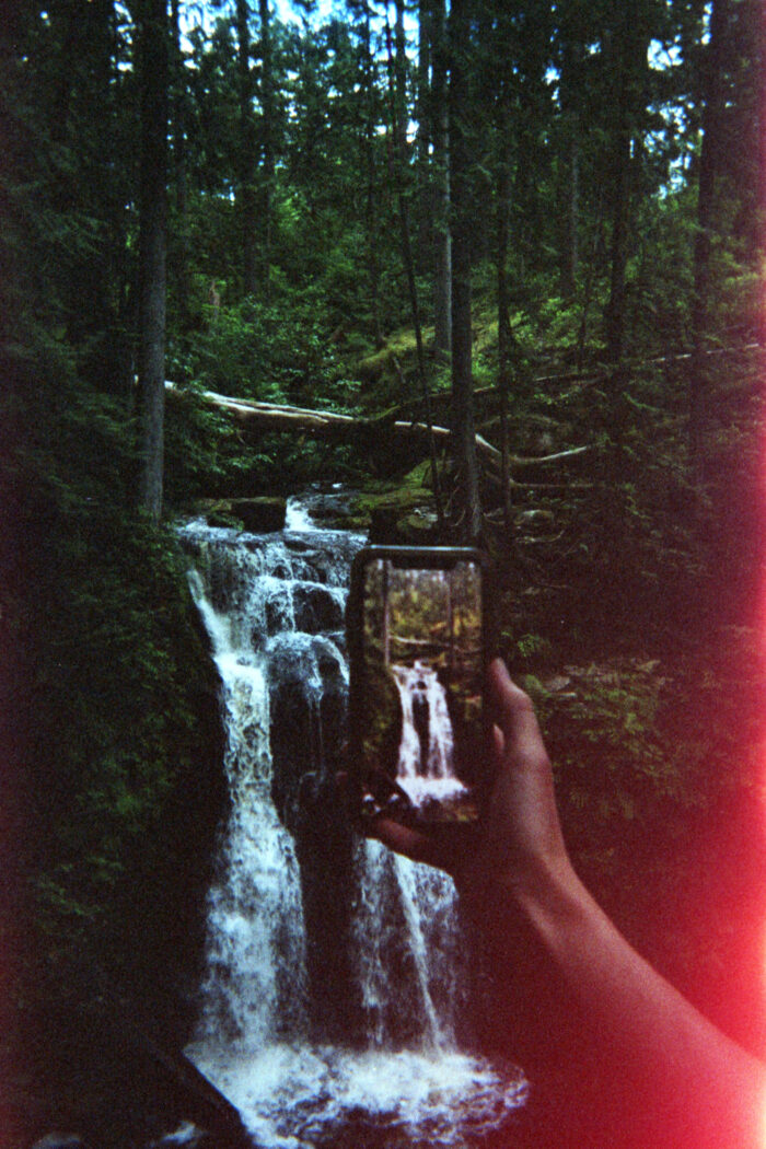 The arm of a light-skinned person is holding an iPhone to photograph a waterfall. There is water flowing down from a rock formation with evergreen trees surrounding the waterfall. There is a red light leak in the photo on the right.