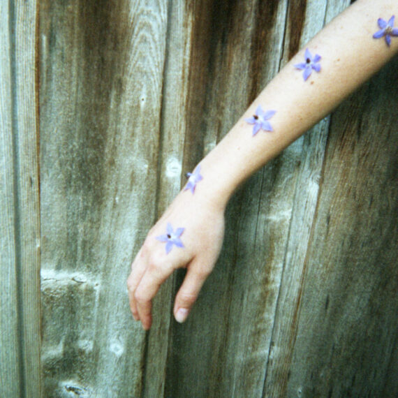 Arm of a pale-skinned person covered in small purple flowers. The person’s arm is outstretched in front of a wooden surface.