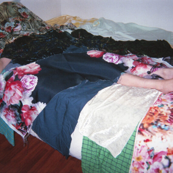 A pale-skinned person lying down on a bed. They are covered with strips of cloth with different colours and patterns. Their whole body is covered in patterned cloth except for one arm and their feet.