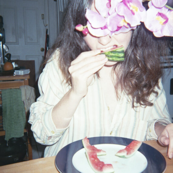 A pale-skinned person with long brown hair is wearing a vertically striped shirt. They are sitting at a wooden table and eating a slice of watermelon. In front of them is a white plate with a black rim holding three watermelon rinds. Orchids are covering the person’s face.