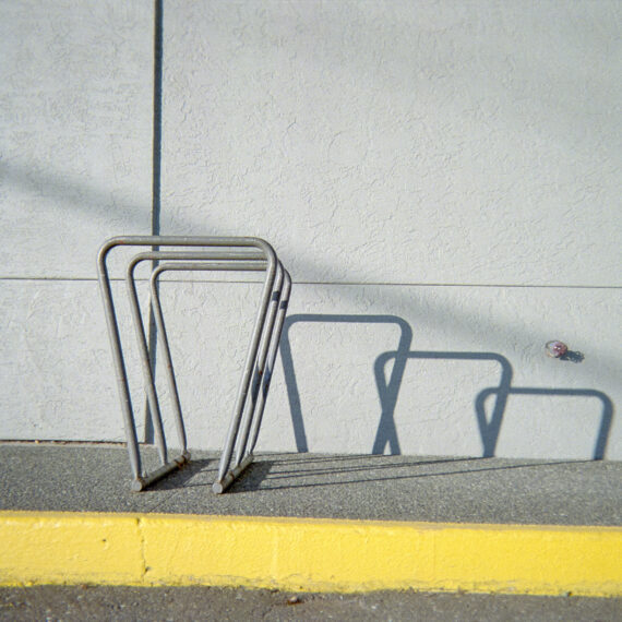 Photo of a bike rack mounted to the pavement. The side of the pavement is painted bright yellow. The sunlight is casting shadows of the bike rack against the wall creating a cascade of shadows of the bike rack shape.