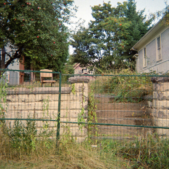 Colour photo of a set of concrete steps with a chair at the top. The property is overgrown with weeds and it is blocked off by a blue fence.