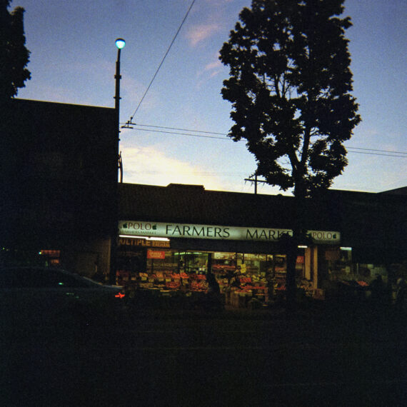 An underexposed photo of a local fruit market that is still open. In ront of it is a tree and a parked car. The sky is blue and purple as the sun sets. Everything around the store is a silhouette while the fruit market is still lit.
