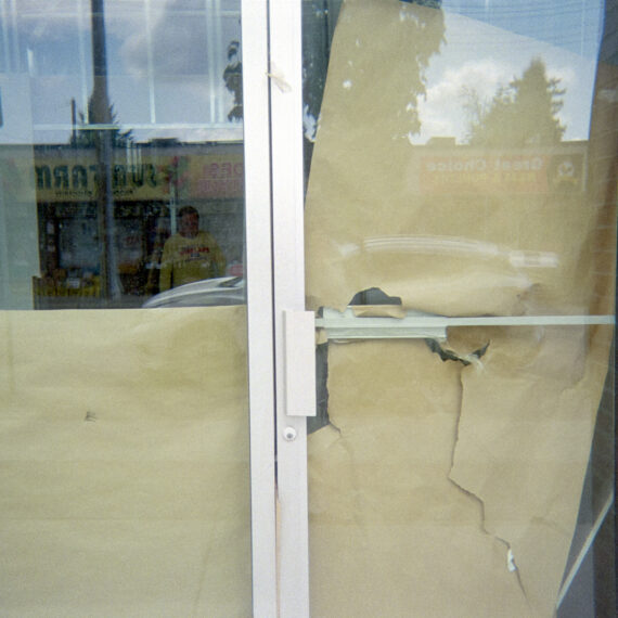 Photo peering into a store front with someone standing inside. The glass doors are partially covered with craft paper that is ripping apart. The window reflection shows the stores across the street.
