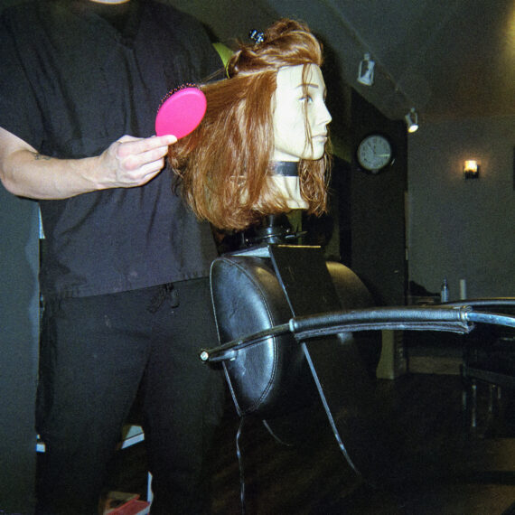 Someone wearing a black shirt and pants blow drying and combing the hair of a mannequin with red hair and white skin. The person is in a salon behind a black chair.