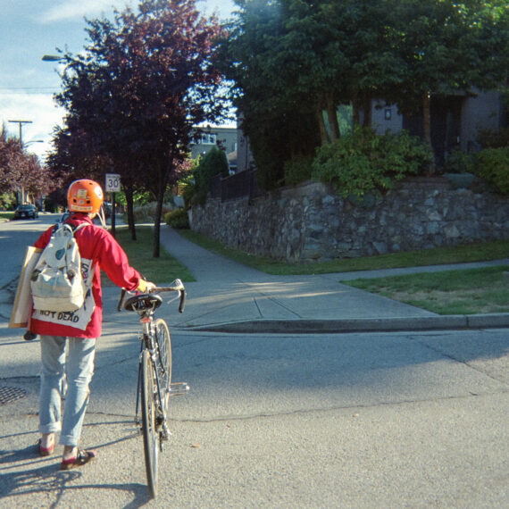 Person wearing a red jacket and an orange helmet walking a bike across a residential street. There is a patch on their jacket partially covered by their backpack that reads "NOT DEAD."