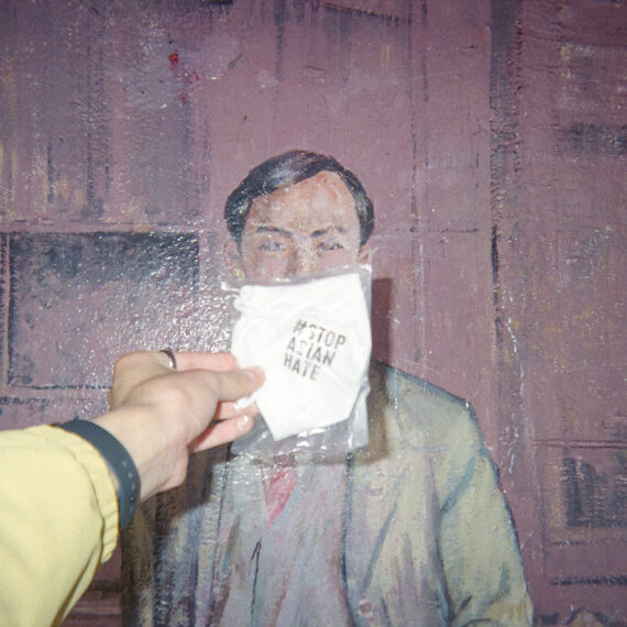Colour photo of a person wearing yellow jacket holding a cloth face mask printed with text that reads "#StopAsianHate." The face mask is being held in front of a historical painting of a Chinese person with short hair in British Columbia.