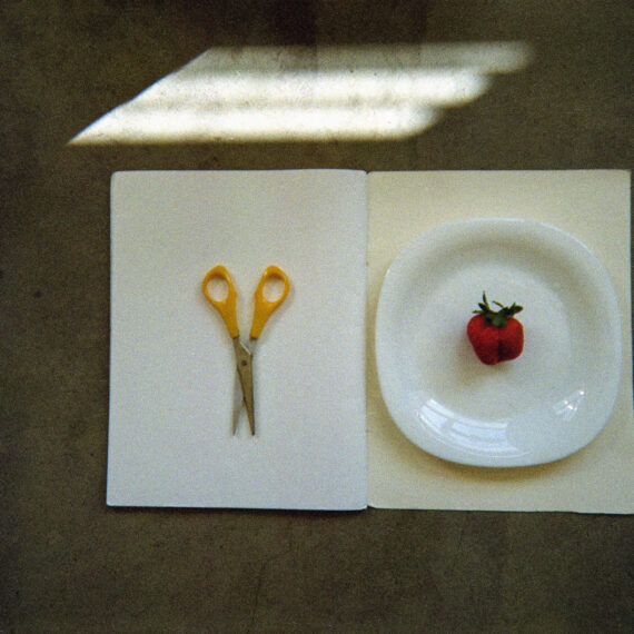 A blank notebook laid flat and open on a concrete surface. There is a yellow pair of scissors and a white plate with a crocheted tomato resting on it. The plate and scissors are resting on top of the open notebook. There are bars of sunlight shining on the floor.
