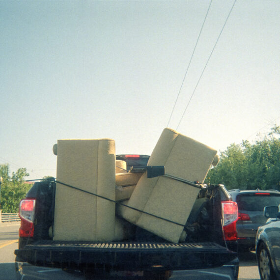 Photo of a pick up truck stopped in traffic. The trunk has two light yellow couches strapped to one another with a black bungee cord. The truck is headed towards a clear blue sky and green trees.