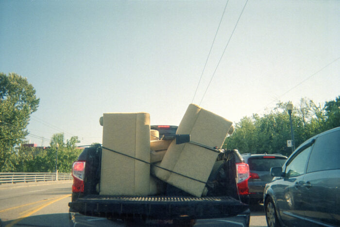 Photo of a pick up truck stopped in traffic. The trunk has two light yellow couches strapped to one another with a black bungee cord. The truck is headed towards a clear blue sky and green trees.