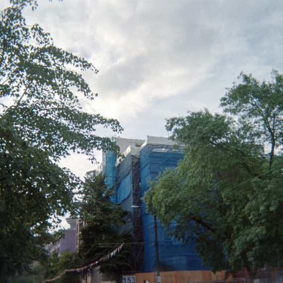 Photo of a building surrounded by blue netting and scaffolding. Surrounding the building are trees against a overcast blue sky.
