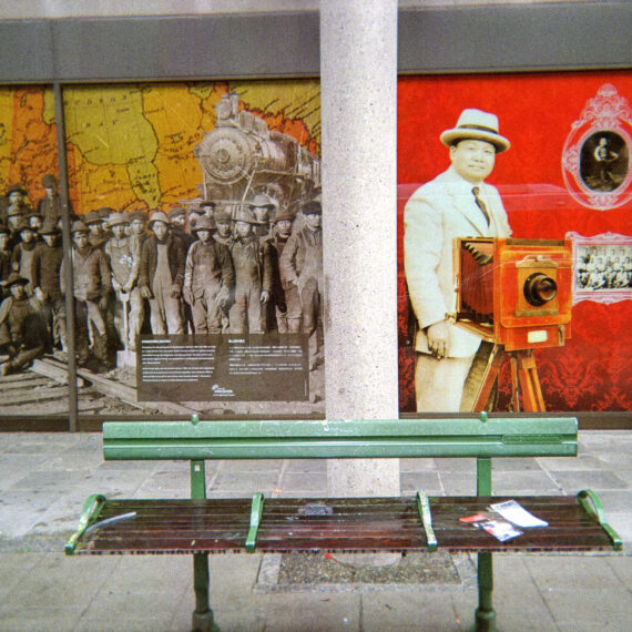 Photo of a bench on the street of Vancouver's Chinatown. Behind the bench is a window display about Yucho Chow, a Chinese Canadian photographer.