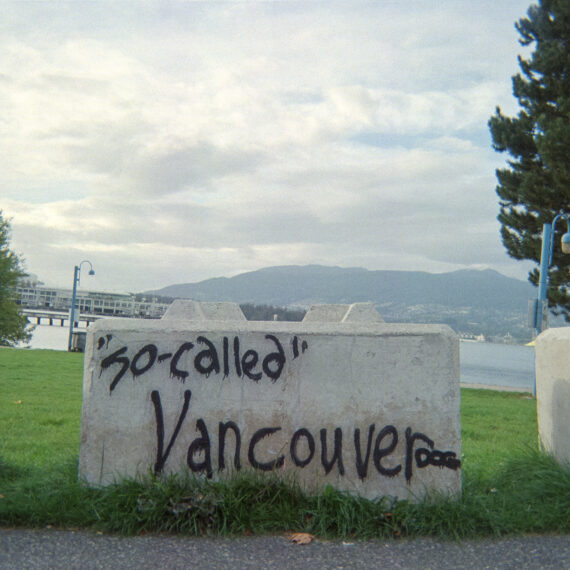 Colour photo of concrete blocks lined up on grass. In the background is a cloudy blue sky with mountains and trees in the distance. One of the concrete blocks is graffitied with black text that reads "so-called Vancouver".