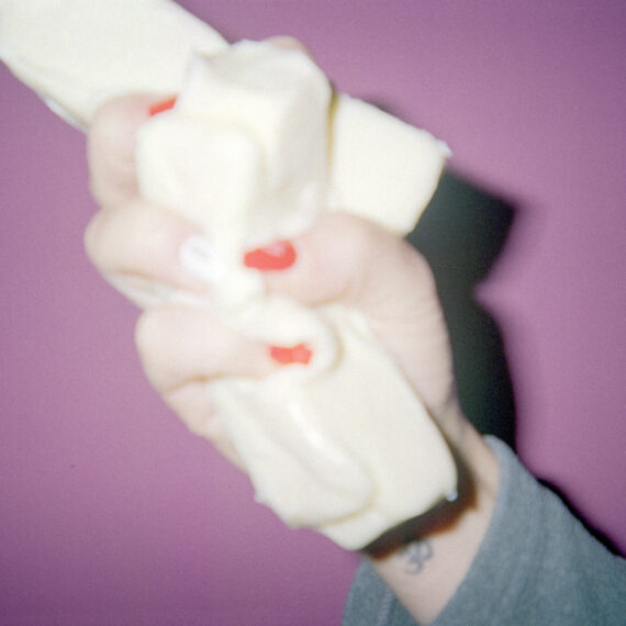 A hand with red painted nails squeezing two sticks of butter. The hand is in front of a magenta background.
