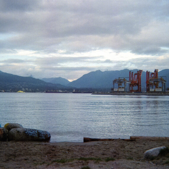 Photo of a beach shore. There is water in the distance as well as red container cranes.