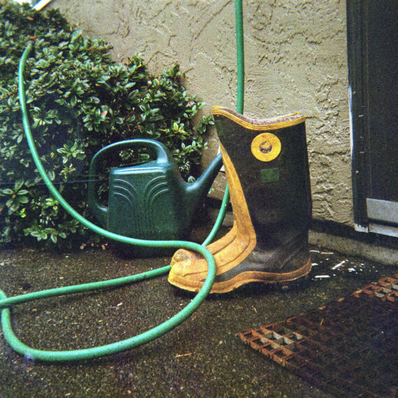 A pair of black and yellow rain boots resting next to a dark green watering can. The boots and water can and resting between a doormat and a shrub.